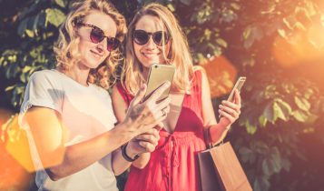 Ein Sommertag, zwei Frauen stehen lächelnd im Park und schauen sich Fotos auf dem Smartphone des anderen an. Die Frauen machen eine Pause nach einer ausgiebigen Shopping Tour